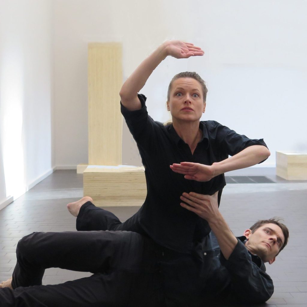 2 dancers dressed in black. One dancer is lying on his back on the floor, balancing the second dancer in a U shape on his side. The balanced dancer has one hand above her face and one below almost making a frame.